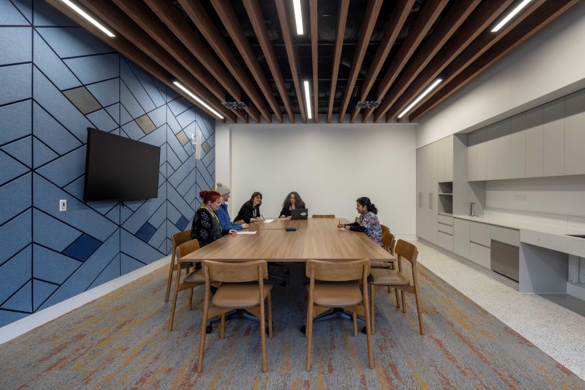 Meeting room with a wooden table and 5 people sitting at the far end, a wall of cupboards on the right side and a TV on the left side.